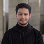 Smiling young man in a black hoodie standing outdoors, exuding confidence and warmth.