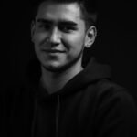 Black and white portrait of a smiling man with earrings and nose piercing, exuding a confident demeanor.