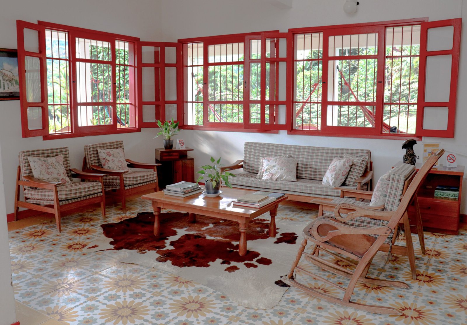 A bright living room with red window frames, wooden furniture, and decorative floor tiles.