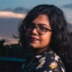 A striking portrait of a woman with eyeglasses at twilight, showcasing a vibrant urban background.