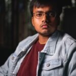 Portrait of a serious young man wearing a denim jacket and eyeglasses indoors.