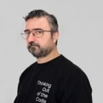 Bearded man wearing eyeglasses and a slogan sweater, looking thoughtful indoors.