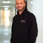 Smiling man in hoodie standing confidently in brightly lit hallway.