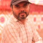 A middle-aged man with a beard and cap poses indoors with a patterned background.