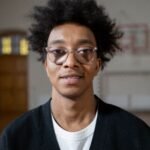 Young man with glasses and afro hair smiling indoors. Casual portrait capturing a relaxed vibe.